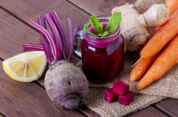 There is a glass of carrot beet ginger juice on the table and next to drink there is ginger, lemon, carrot and ginger 